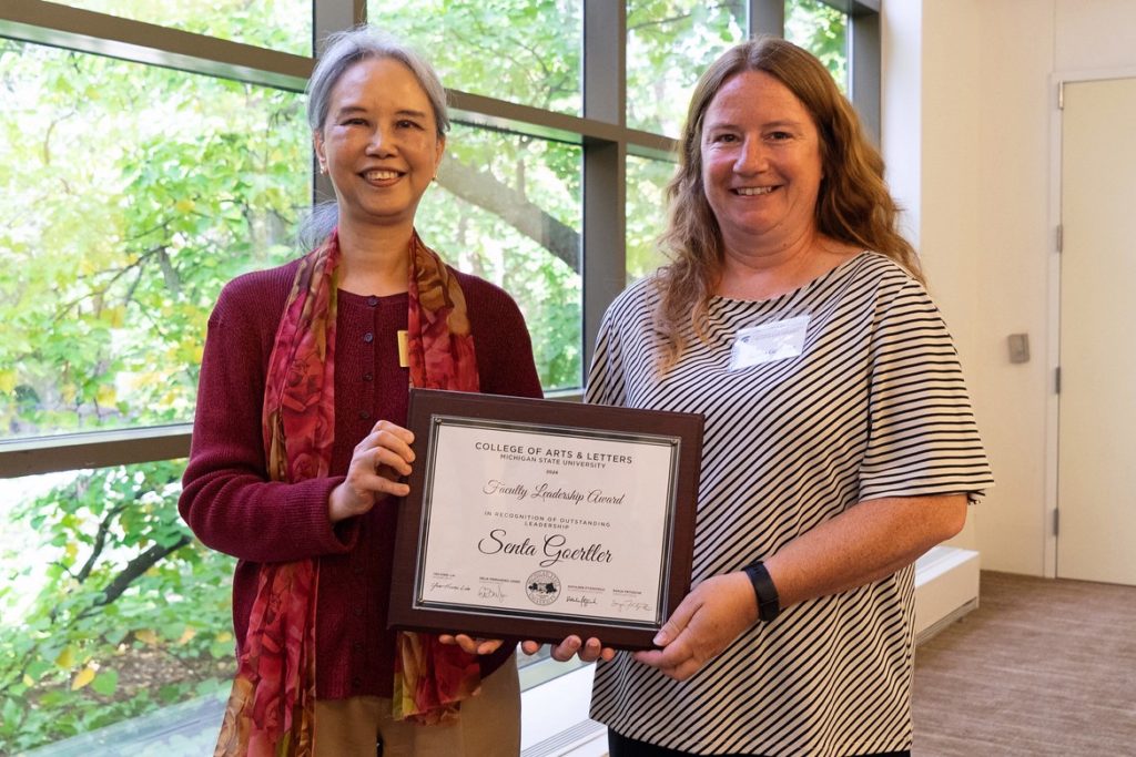 Two women stand smiling and holding an award certificate. The woman on the left wears a red sweater and scarf, and the woman on the right wears a striped top. They stand in front of a large window with greenery visible outside. 